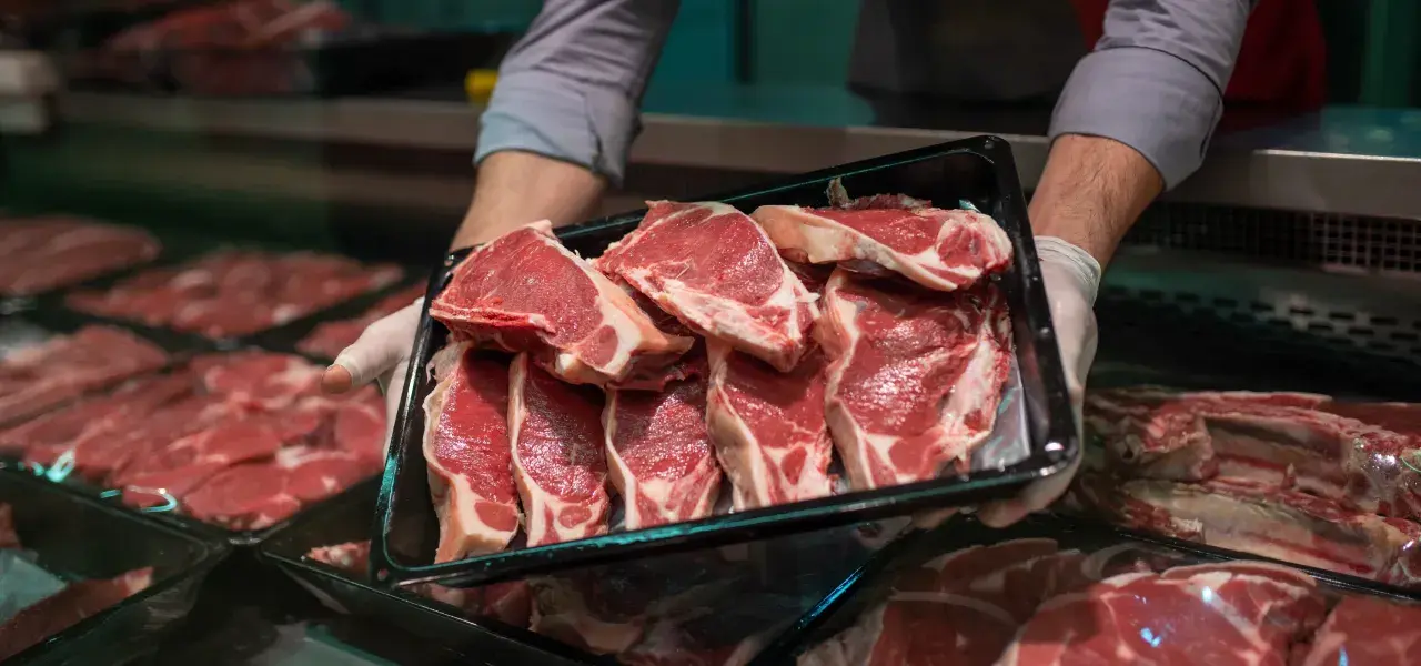 Butcher holding a tray of fresh lamb chops at a meat counter.