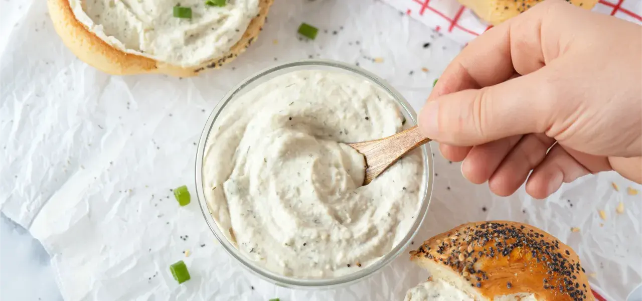 Hand stirring creamy herb cheese spread in a glass bowl next to bagels.
