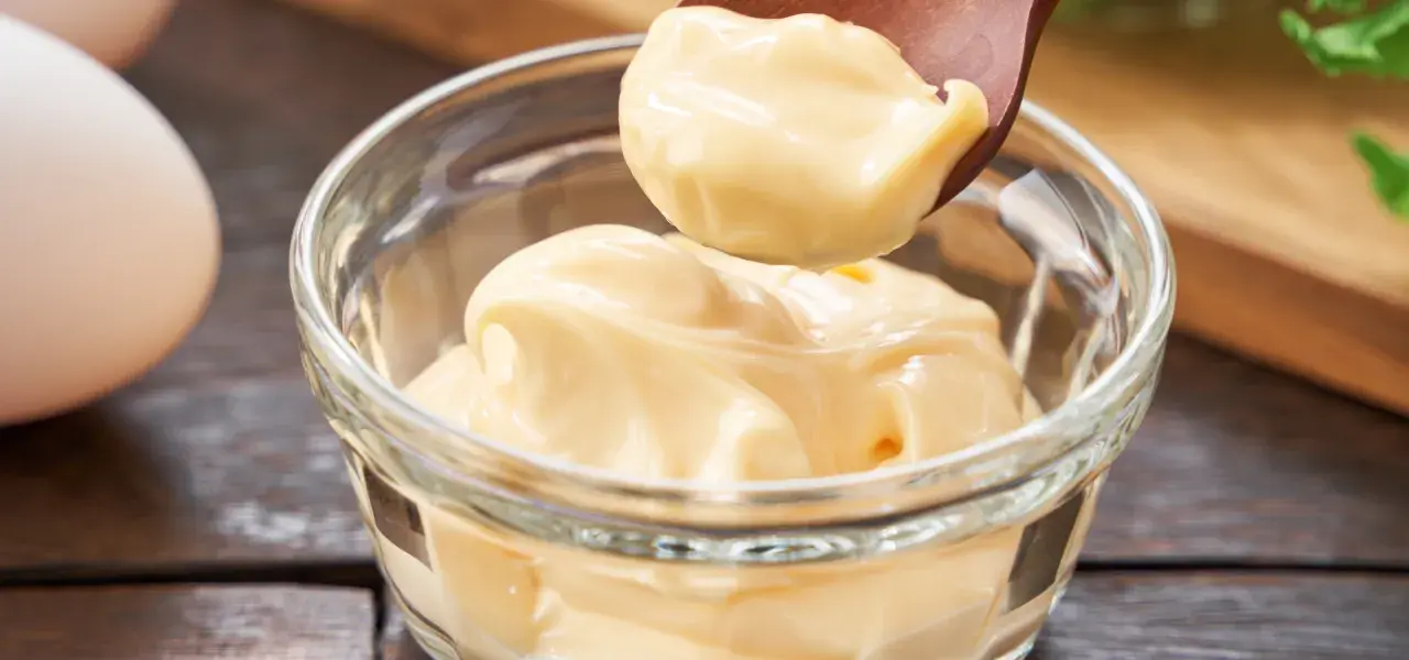 Wooden spoon scooping creamy mayonnaise from a glass bowl.
