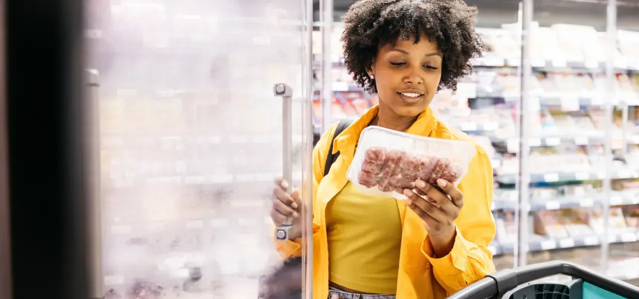 Woman shopping for packaged meat in a grocery store freezer aisle.