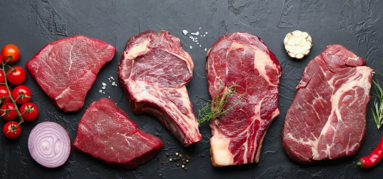 Assorted raw beef cuts with vegetables and spices on a dark surface.